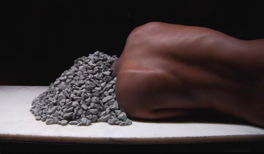 A pile of small gray stones next to a clenched human fist on a white surface against a dark background.
