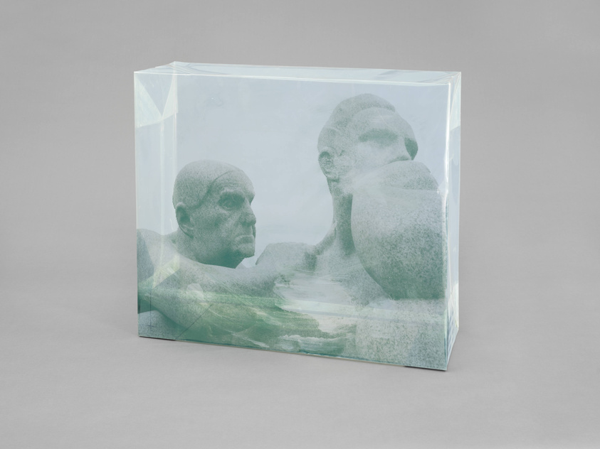 A sculpture of two human figures encased in a transparent block, giving the appearance of being frozen within ice or suspended in clear resin. The figures are detailed and appear contemplative, with one facing outward and the other with its head turned to the side. The sculpture is set against a neutral gray background.