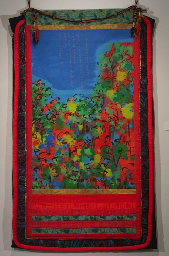 A vibrant textile artwork hangs on a wall, featuring a central blue panel with vertical lines of text, surrounded by a colorful depiction of trees and foliage in red, green, yellow, and orange hues. The piece is bordered by a red frame with intricate patterns and tassels at the top corners.