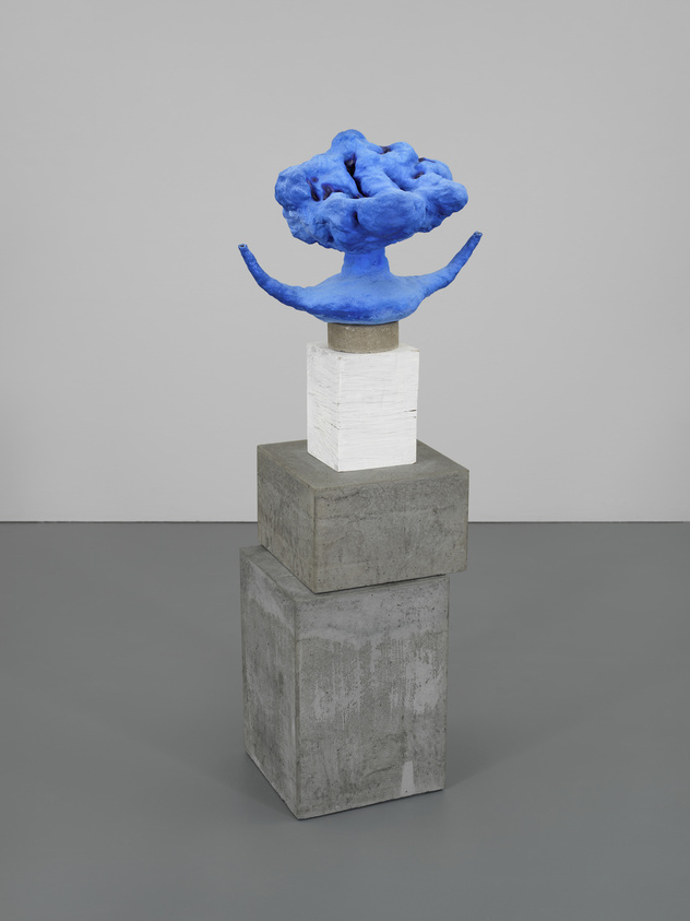 A vibrant blue abstract sculpture with undulating forms and protrusions sits atop a white and gray pedestal against a neutral background.
