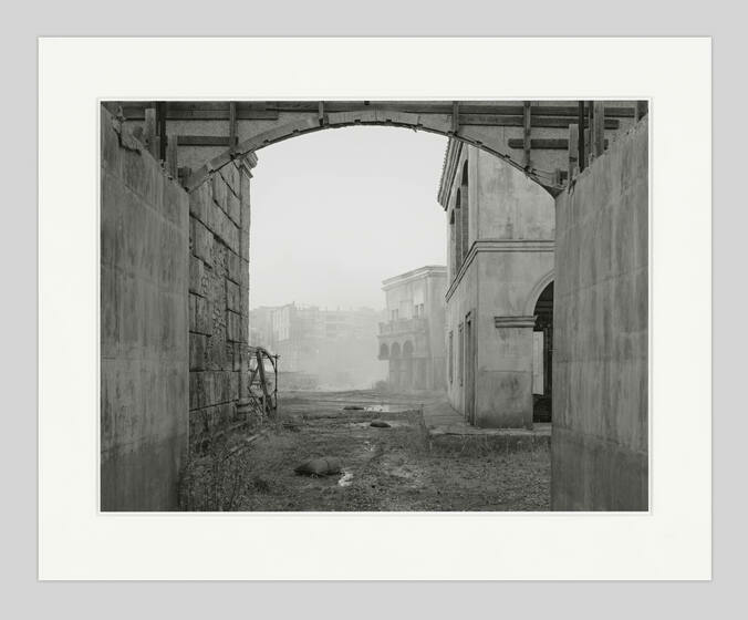 Black and white photograph of a misty scene viewed through an archway, showing dilapidated buildings and debris, with a sense of abandonment and decay.