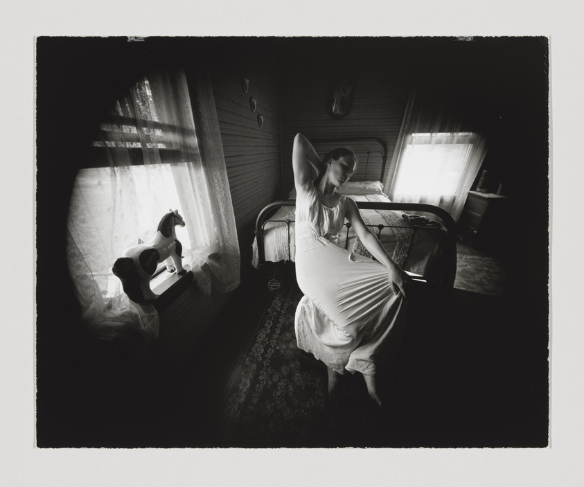 A black and white photograph capturing a woman in a vintage dress sitting on the edge of a bed in a room with light filtering through sheer curtains. The room has a nostalgic feel with a rocking horse visible on the left and a wall-mounted animal head above the bed. The woman appears to be in a contemplative or relaxed pose, with one hand behind her head and the other resting on her knee. The image has a dreamlike quality due to the soft lighting and wide-angle perspective.