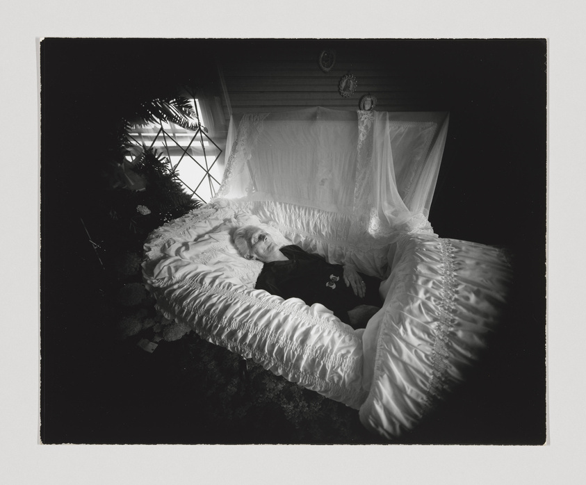 A black and white photograph depicting an elderly person lying in an ornate coffin with a satin interior. The coffin is placed in a room with a window in the background, allowing natural light to illuminate the scene. The person is dressed in dark clothing with a brooch, and the coffin is adorned with sheer drapery. The overall atmosphere of the image is somber and reflective.