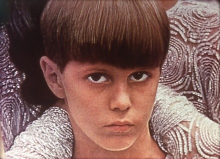 A close-up of a young person with a bowl haircut, looking directly at the camera with a serious expression. They are wearing a garment with a detailed, sequined pattern.