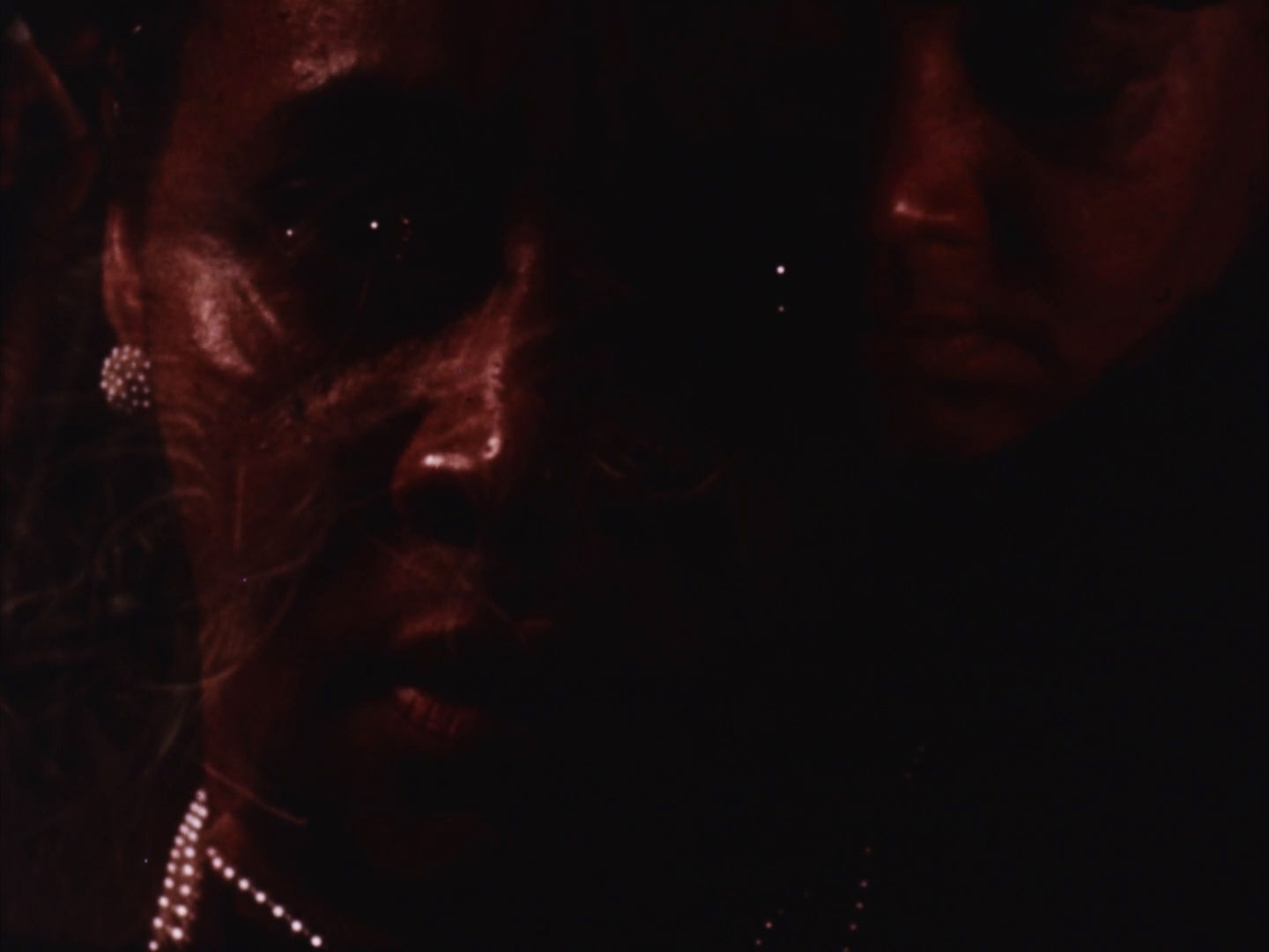 A close-up, low-light image of two people's faces, with the focus on the person in the foreground who has visible pearl earrings and a necklace. The image has a reddish hue, adding to the dramatic and moody atmosphere.