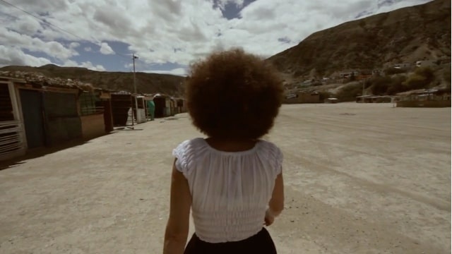 Person with afro walking down a dusty road in a mountainous area with sparse buildings under a cloudy sky.