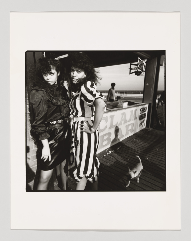 Two women posing confidently on a boardwalk with a food stand in the background advertising clam bar and shish kebab. The woman on the left is wearing a black leather jacket and skirt, while the woman on the right is in a black and white striped dress. A small dog is seen walking by in the foreground, and another person is visible in the distance against a bright sky. The photo has a vintage feel, captured in black and white.