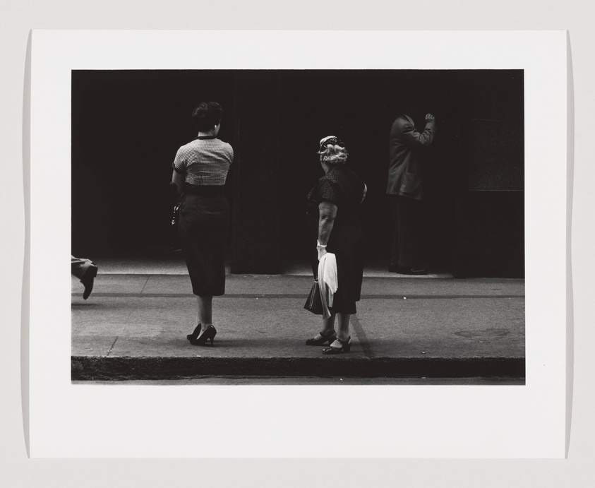 A black and white photograph capturing a street scene with three individuals: a woman in a striped top and skirt walking to the left, an older woman standing in the center facing right, and a man in a suit leaning against a dark wall in the background. The contrast between light and shadow is stark, highlighting the figures against a mostly dark backdrop.