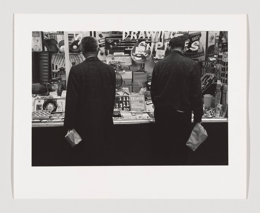 Two men are standing in front of a store window filled with various items, including drawing supplies and cameras. The man on the left is wearing a dark suit and holding a small bag, while the man on the right is dressed in a dark jacket with a pattern and also holding a bag. They appear to be looking at the items displayed in the window. The photo is black and white, capturing a moment of everyday life, possibly suggesting a scene of urban window-shopping.