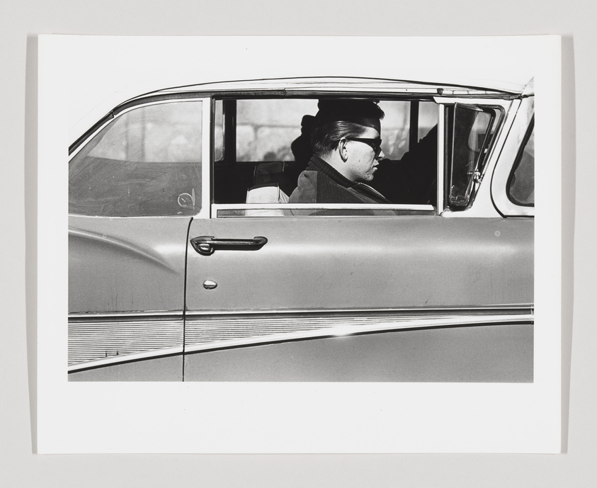 A black and white photograph capturing a side view of a person wearing sunglasses and a hat, sitting in the driver's seat of a vintage car with the window partially rolled down. The car's exterior reflects a sleek mid-century design, with a prominent side trim and a door handle visible. The person appears focused, possibly on the road ahead, and the image is framed to include only the car's side profile and the driver.