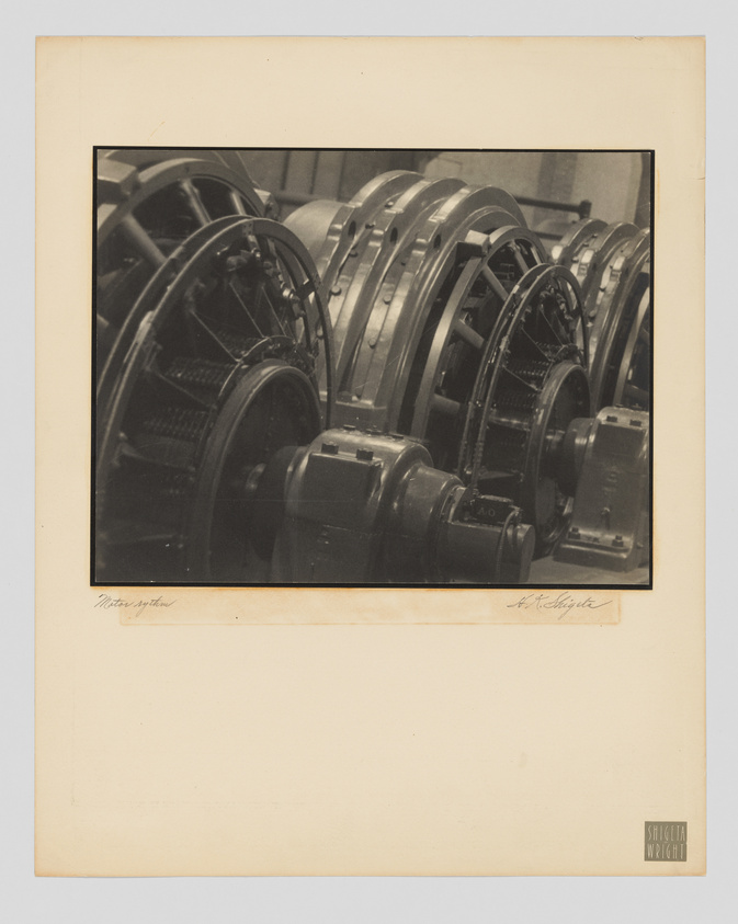 A sepia-toned photograph depicting a series of large, industrial metal wheels, possibly part of a machine or engine, with intricate spokes and gears visible. The image is mounted on a beige card with handwritten text at the bottom left and a signature at the bottom right, along with a stamp of the photographer's studio name, "Shigeta Wright."