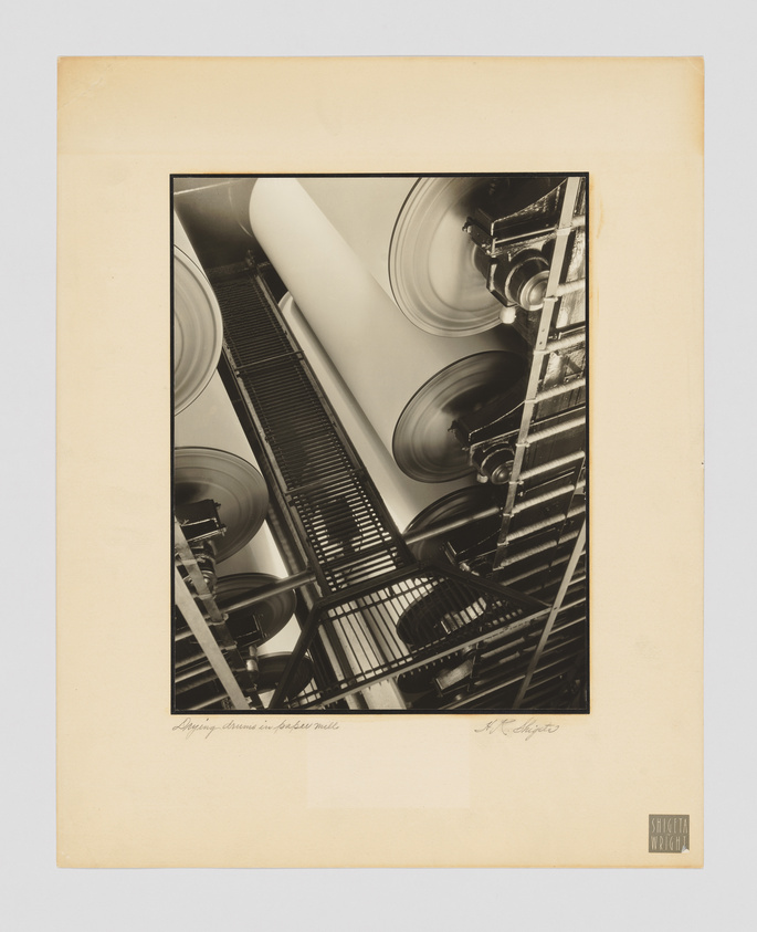 A black and white photograph mounted on a beige mat board, depicting an upward view of a building's industrial features, including large cylindrical structures and a metal staircase. The image has a dynamic composition with strong lines and contrasts, emphasizing the architectural details. There is handwriting at the bottom of the mat board, likely indicating the title of the work and the photographer's signature.