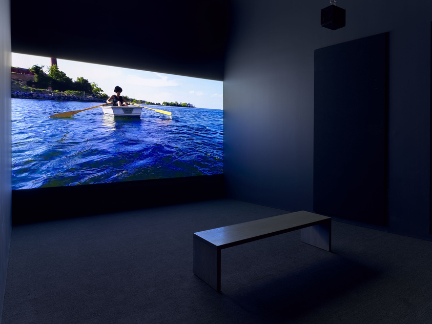 A large projection of a person rowing a boat on a body of water is displayed on a wall in a dark room with a bench in the foreground for viewers to sit and watch.