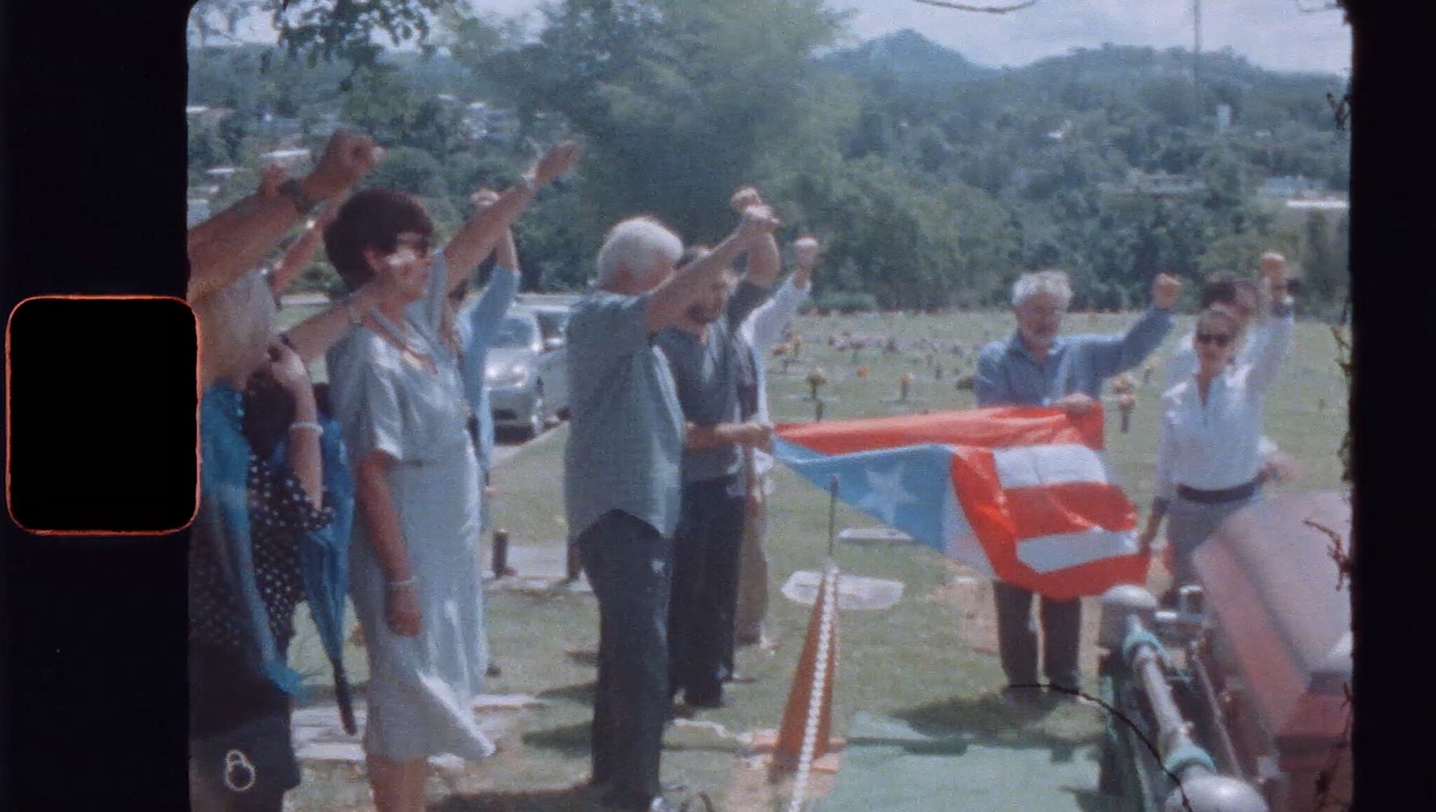 A group of people standing outdoors with their fists raised in a gesture of solidarity or protest. One individual is holding a Puerto Rican flag. The setting appears to be a cemetery with numerous headstones in the background, and the image has a vintage, grainy quality, suggesting it may be from an older film or photograph.