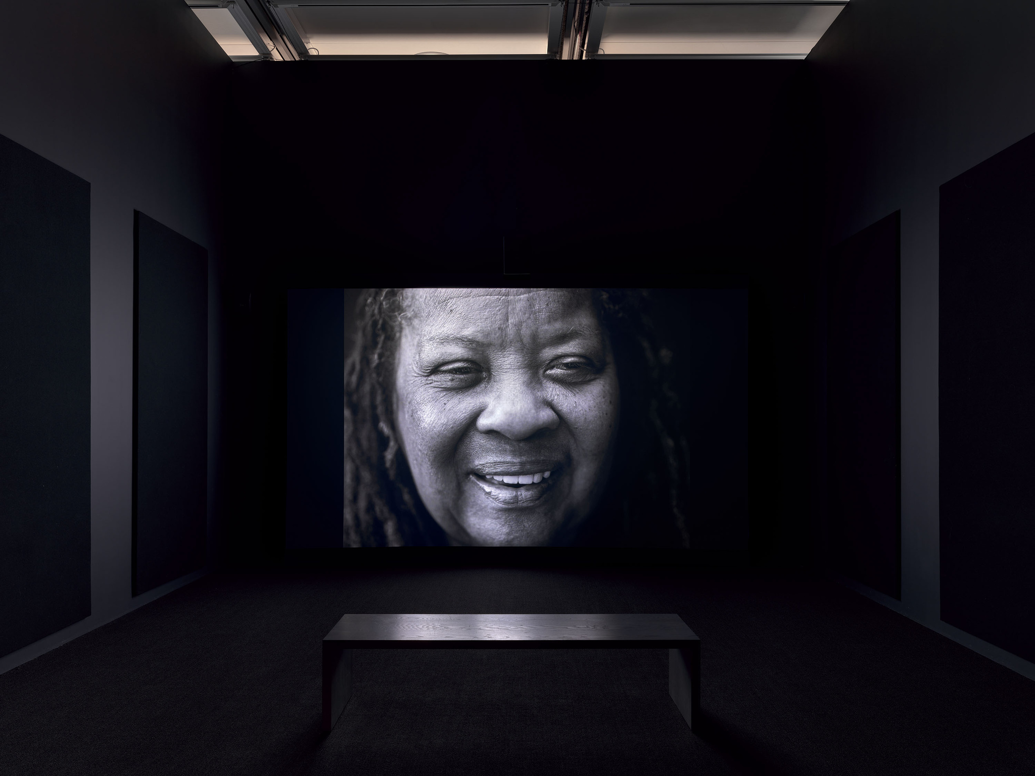 A dark room with a large screen displaying a close-up image of a smiling person with dreadlocks. A bench is placed in front of the screen for viewers.