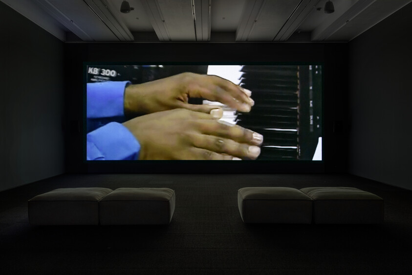 Large screen shows hands playing a keyboard, with two benches in a dark room.