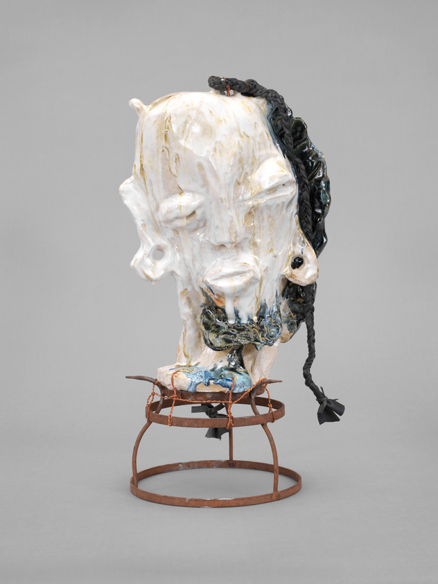 Sculpture of a distorted human head with closed eyes, braided hair, and a textured surface, mounted on a rusted metal stand.
