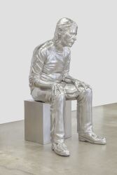 A shiny, metallic sculpture of a seated person with a contemplative pose on a square pedestal in a gallery setting.