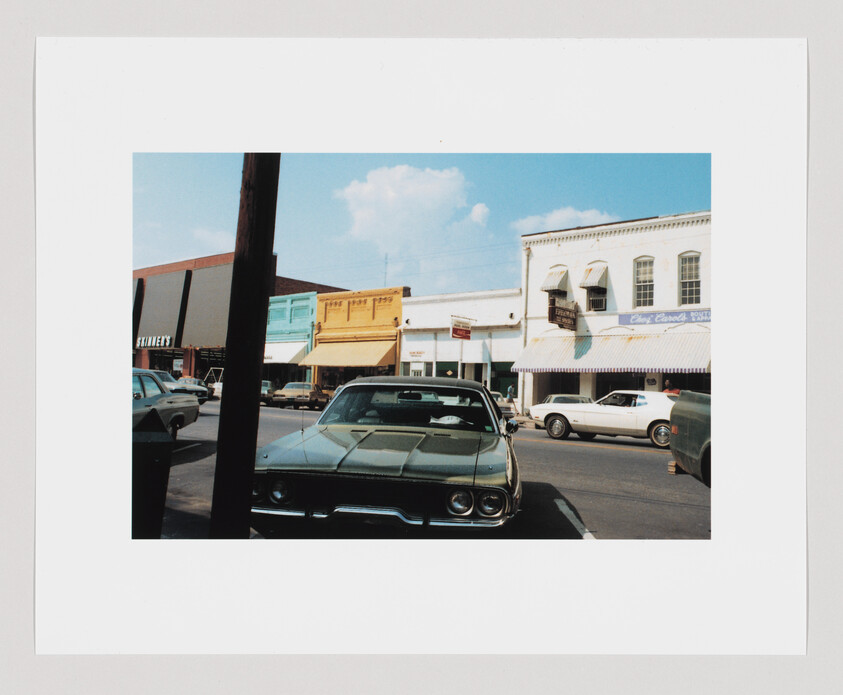 A green vintage car is parked on a street with several shops and other cars in the background.