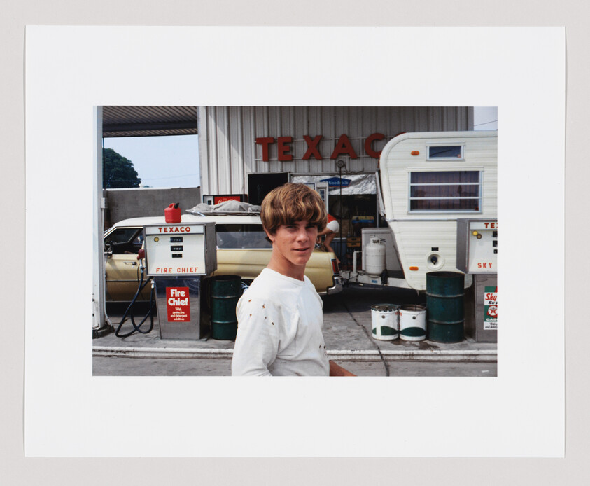 Teenage boy with light brown hair stands in front of a Texaco gas station, next to pumps and a trailer.