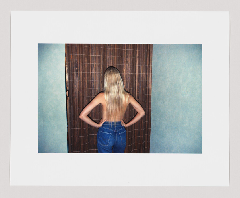 A person with long blonde hair, wearing blue jeans, stands facing a brown curtain with hands on hips.