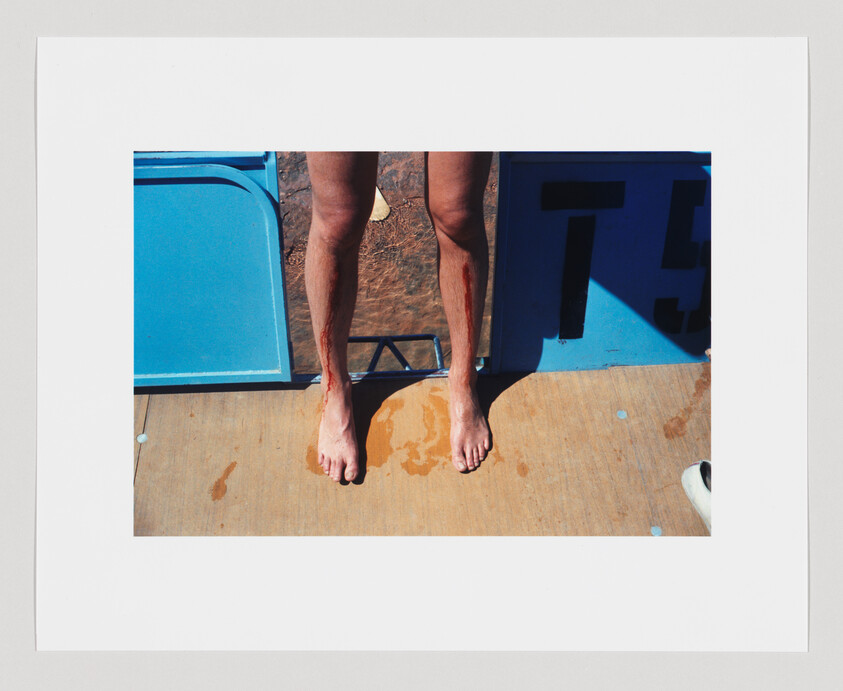 Bare legs and feet with streaks of blood standing on a wet surface near a blue platform marked “5.”