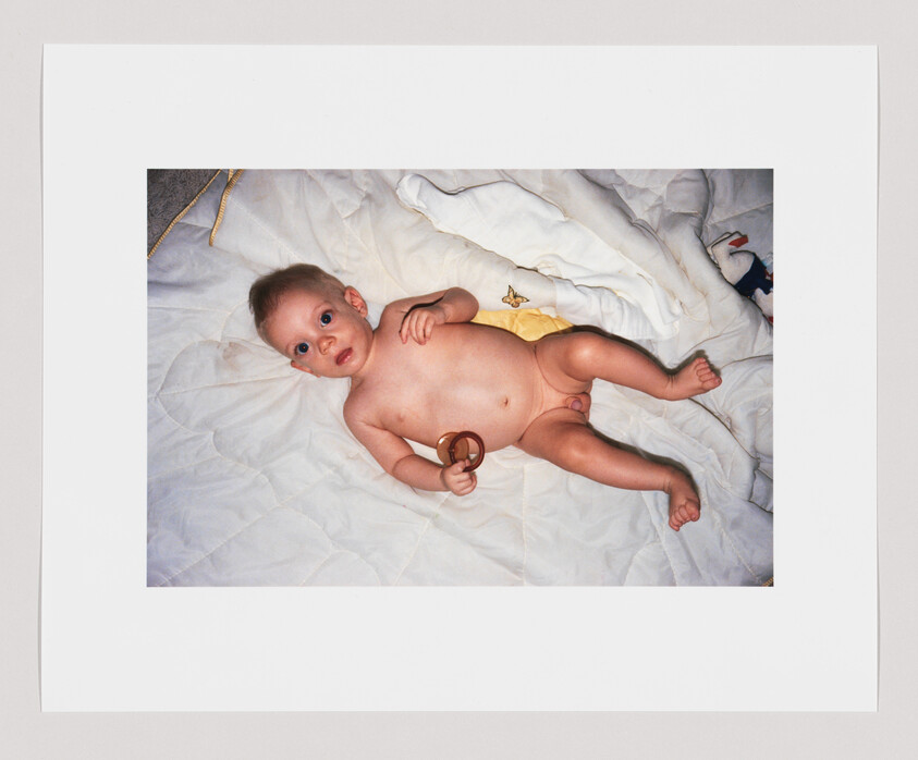 A baby lying on a white blanket, holding a pacifier, looking up with wide eyes.