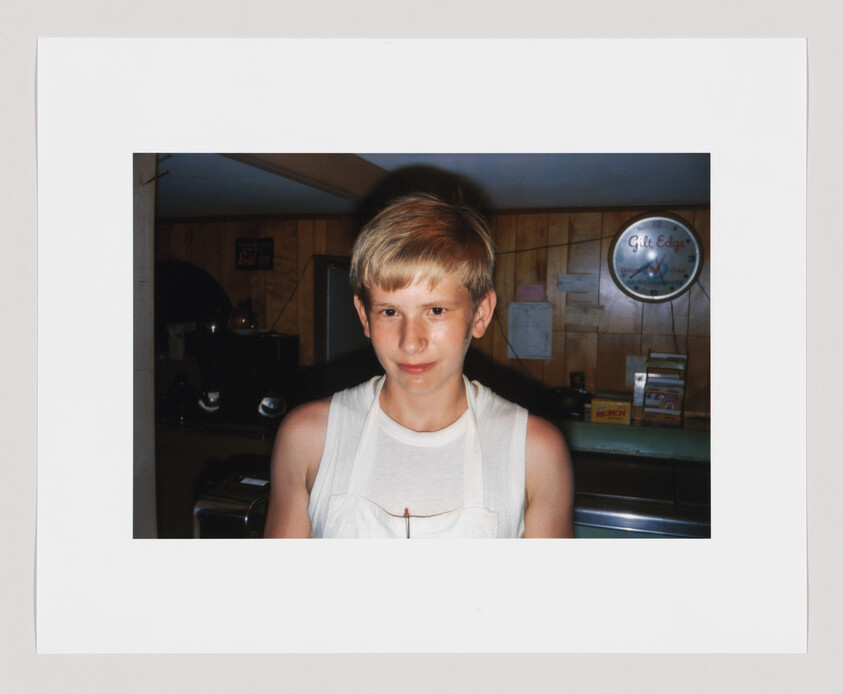 Young boy with blond hair wearing a white apron stands indoors, with a clock and boxes in the background.
