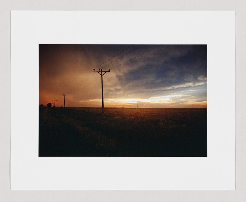 Power lines stretch across a flat field at sunset, with dark clouds and orange light in the sky.