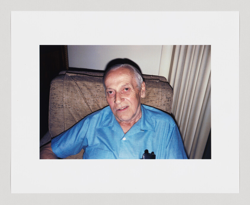 Older man with short gray hair sits on a beige chair, wearing a light blue shirt with a pen in pocket.