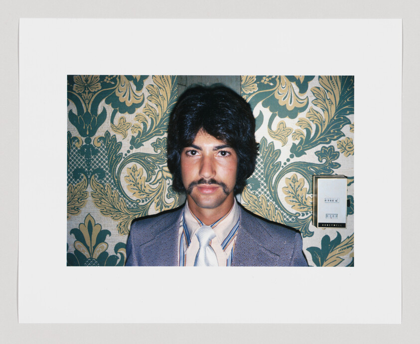 Man with dark hair and mustache wearing a suit and tie, standing in front of patterned wallpaper.