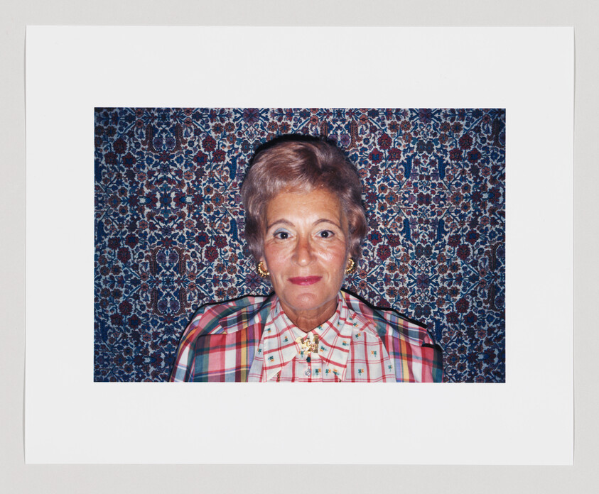 Older woman with short hair and plaid shirt sits in front of a patterned, colorful tapestry background.