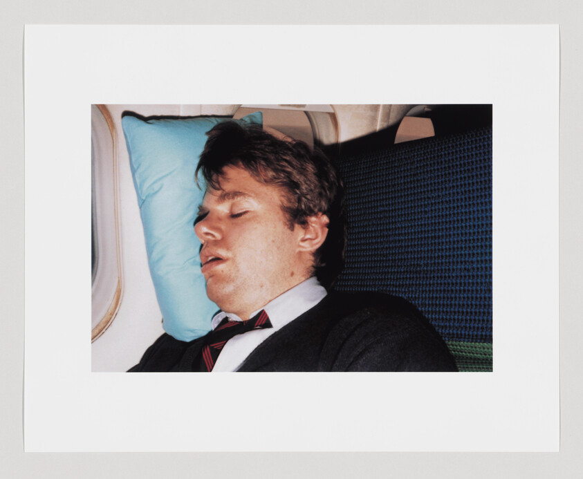 A man in a suit and tie is sleeping with his head on a blue pillow next to an airplane window.