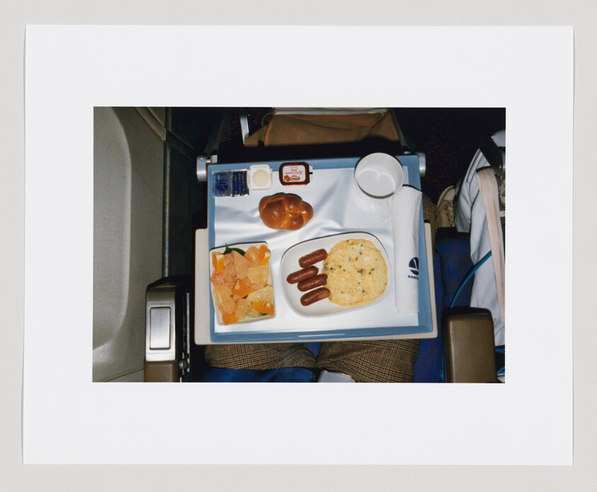 Airplane meal tray with scrambled eggs, sausages, fruit, bread roll, butter, jam, and a cup.