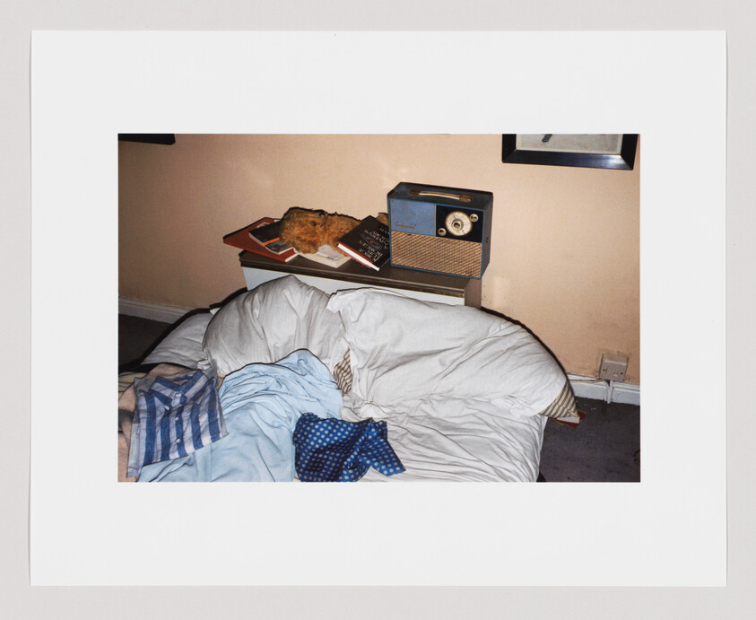 Unmade bed with white pillows, blue blanket, and a nightstand holding books, a radio, and a stuffed animal.