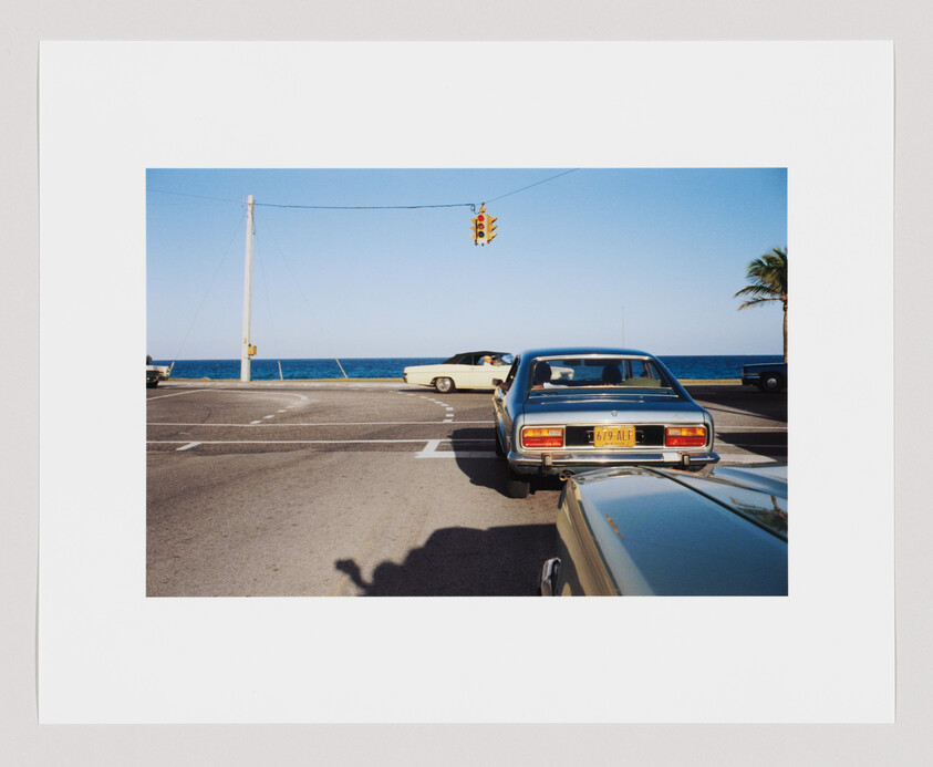 A blue car waits at a stoplight near the ocean, with palm trees and other cars nearby.