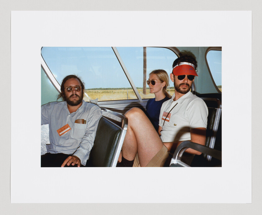 Three adults wearing sunglasses sit quietly on a bus; two have visible name tags on their shirts.
