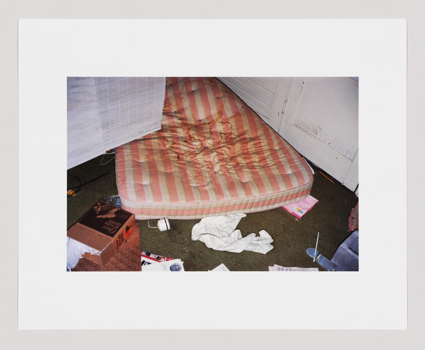 A worn striped mattress on the floor surrounded by scattered clothes, boxes, papers, and a cup.