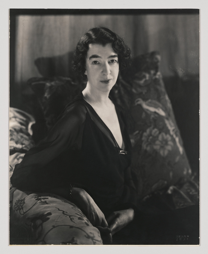 A black and white portrait of a seated woman with dark hair styled in waves, wearing a dark blouse with a V-neckline and a light-colored undergarment. She has a calm expression and is looking directly at the camera. The background features a patterned curtain, and she is resting her arm on a floral upholstered piece of furniture. The photograph has a vintage quality, indicated by the style of dress and the monochrome tones.