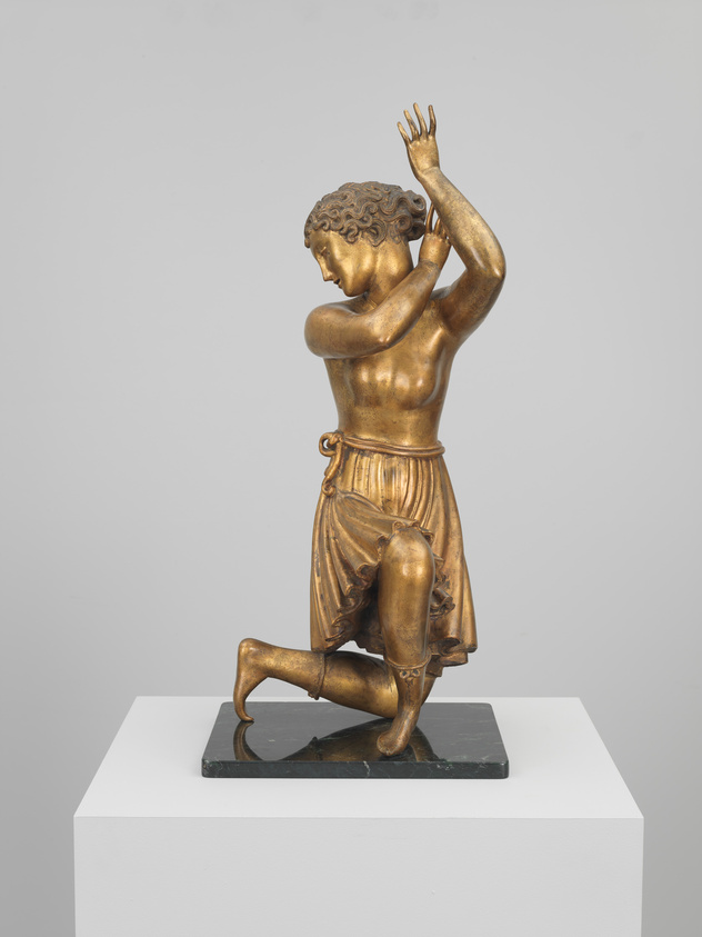 A bronze statue of a kneeling figure with one arm raised, displayed on a white pedestal against a neutral background. The figure has detailed curly hair, is wearing a draped garment around the lower body, and has a muscular build, suggesting a classical or mythological subject.