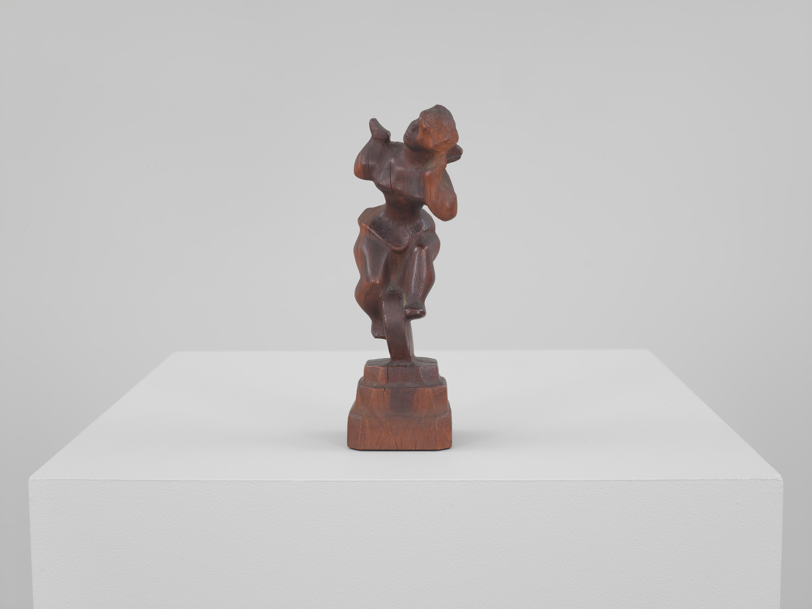 A wooden abstract sculpture displayed on a white pedestal against a neutral background. The sculpture features interlocking forms and curves, suggesting a stylized figure or arrangement of shapes.