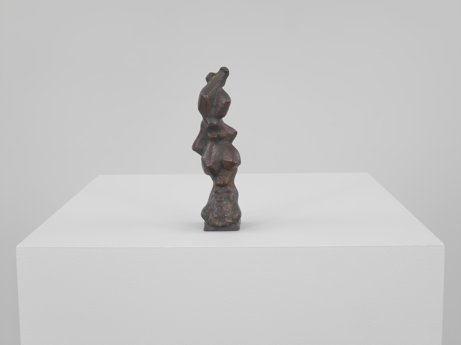 A small, abstract bronze sculpture with an irregular, totem-like shape is displayed on a white pedestal against a neutral background.