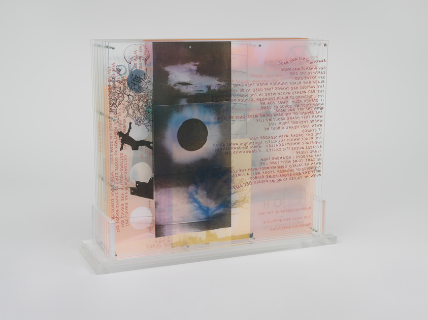 A transparent acrylic sculpture with layered images and text, featuring a mix of celestial motifs, human silhouettes, and various patterns, creating a collage-like effect.