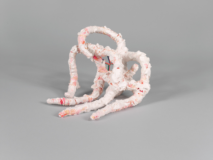 A sculptural artwork resembling a tangle of organic shapes and tubes, predominantly white with hints of pink and red, displayed against a neutral gray background.