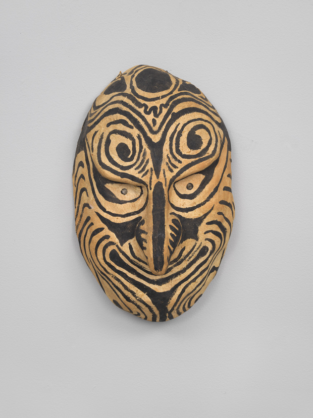 A traditional wooden mask with intricate black patterns on a beige background, displayed on a white wall. The mask features stylized eyes, a nose, and a mouth, with swirling and geometric designs that suggest tribal or cultural significance.