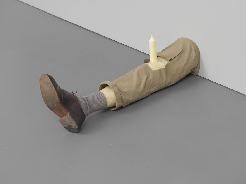 A sculpture of a human leg wearing a brown shoe and grey sock, protruding from a beige fabric sleeve with a candle placed on top, set against a grey floor and white wall.