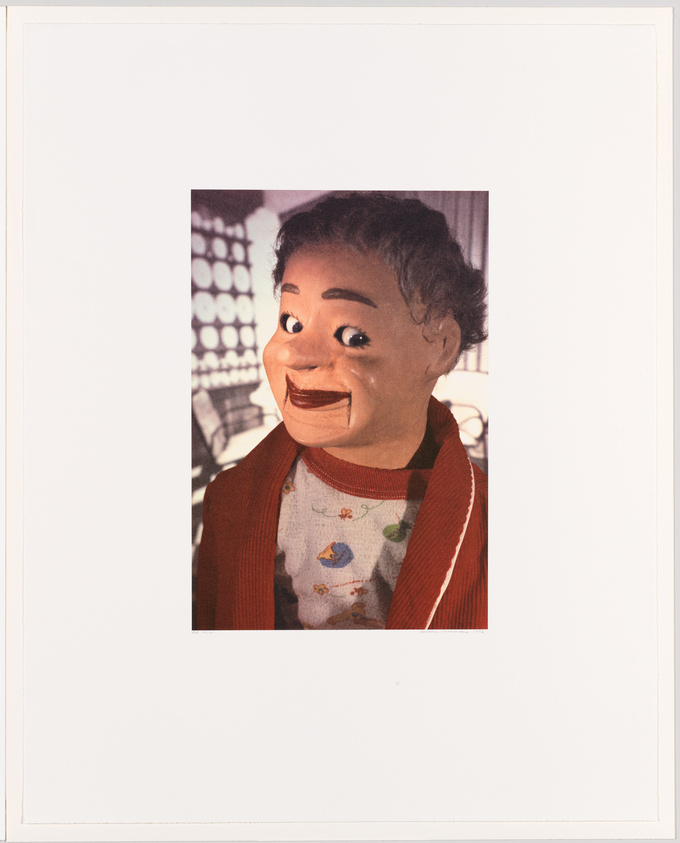 A puppet with a human-like appearance, featuring gray hair and a wide smile, wearing a red cardigan over a shirt with a pattern of flowers and a bird. The background is partially visible, showing a blurred control panel with various buttons and screens.