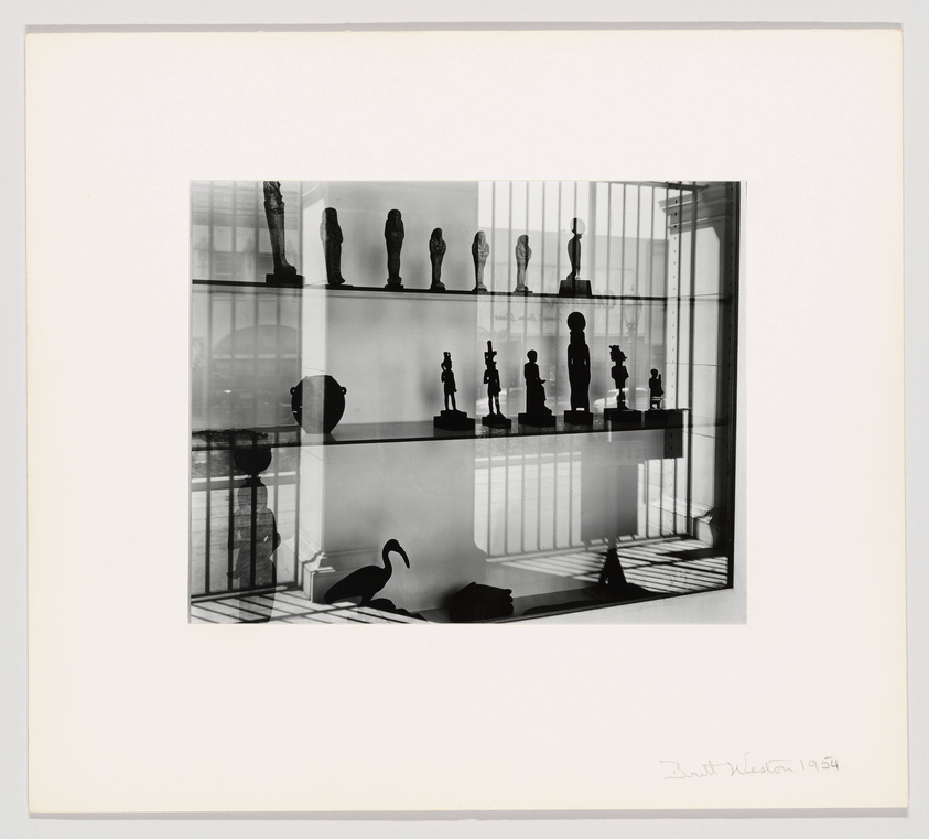 A black and white photograph of various sculptures displayed on shelves with their shadows casting on the surfaces and background, featuring a mix of figurative and abstract forms. The image includes a signature and date in the lower right corner, indicating it was created in 1954.