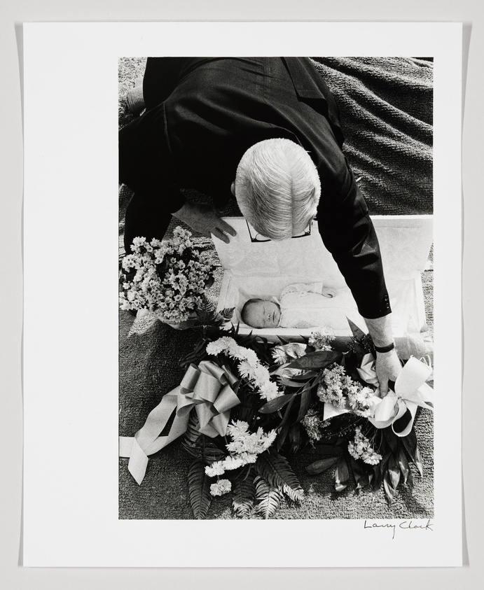 A black and white photograph of an elderly man bending over to look at a sleeping baby surrounded by flowers on a blanket. The image is signed by Larry Clark.