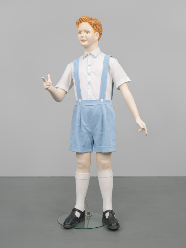 A lifelike statue of a boy in vintage clothing giving a peace sign, standing against a grey background.